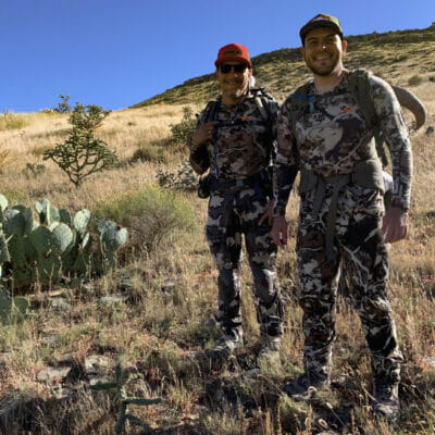 Two men wearing camoflauge smile on a mountain hill.