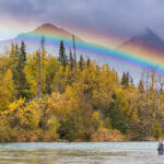 Distant view of a man fishing in a deep river with a rainbow and mountain in the background.